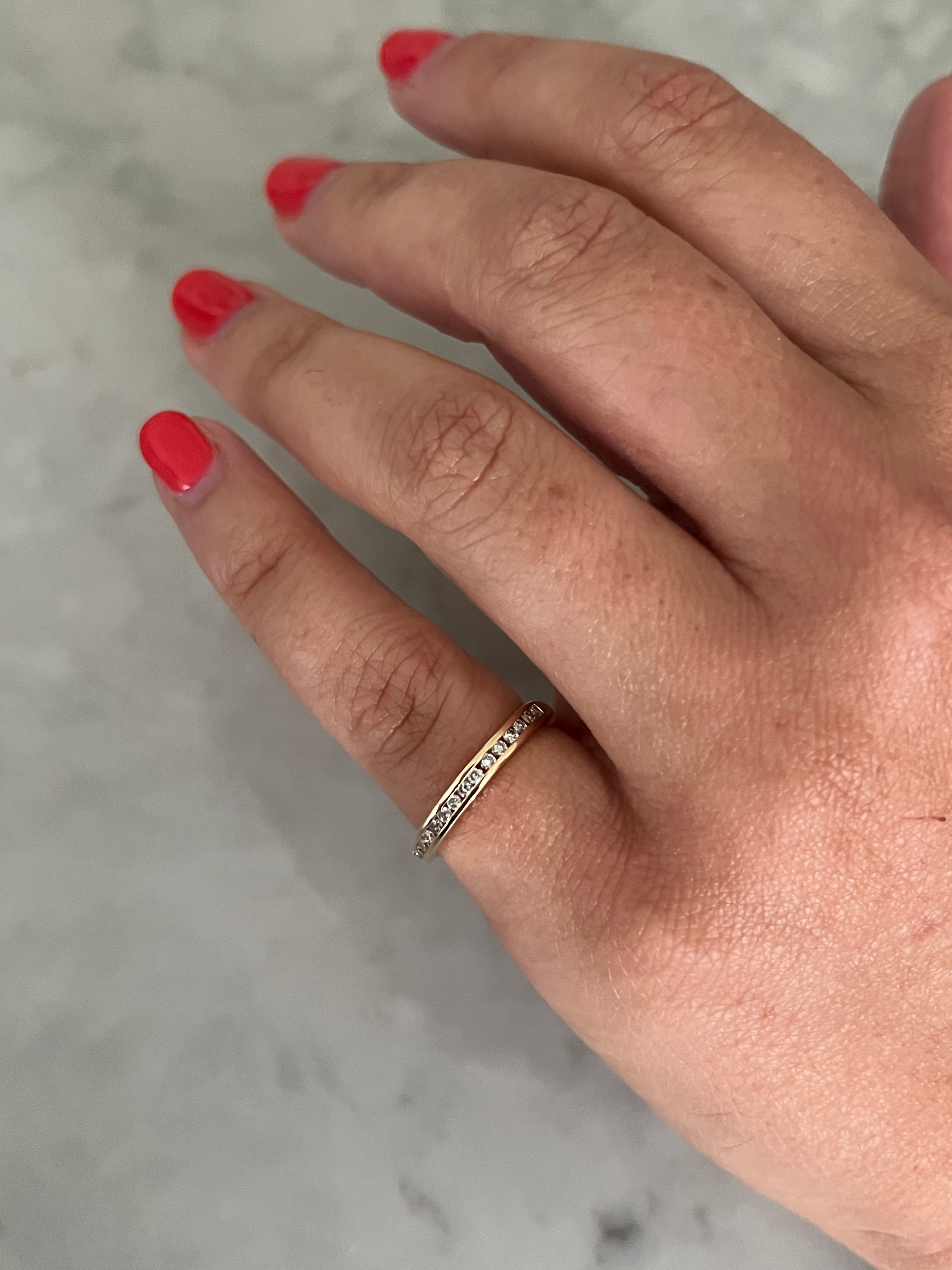 A close-up of a hand wearing a delicate gold and diamond channel-set ring, set against a light gray marble background.