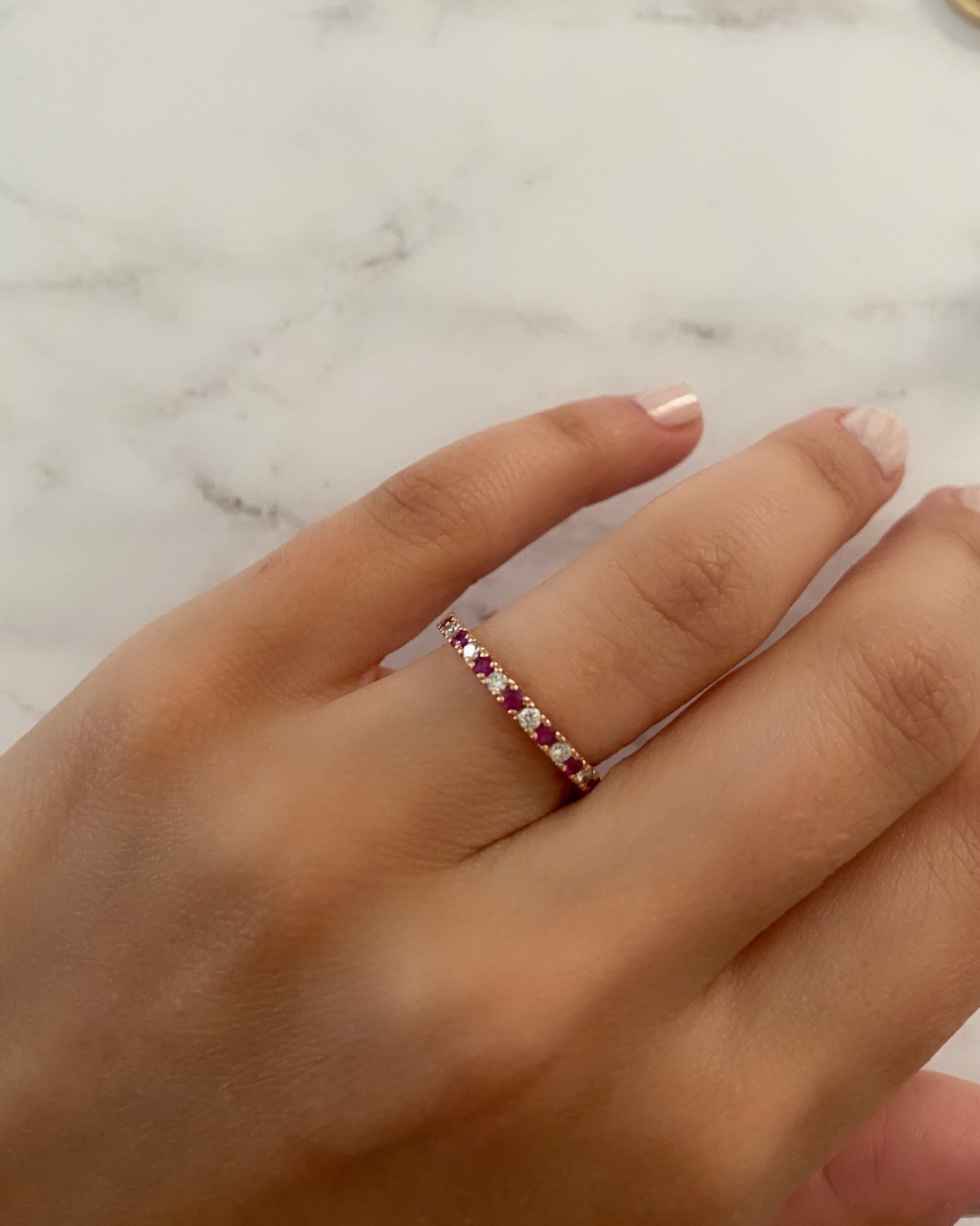 A person's hand wearing an alternating diamond and birthstone ring against a neutral background.