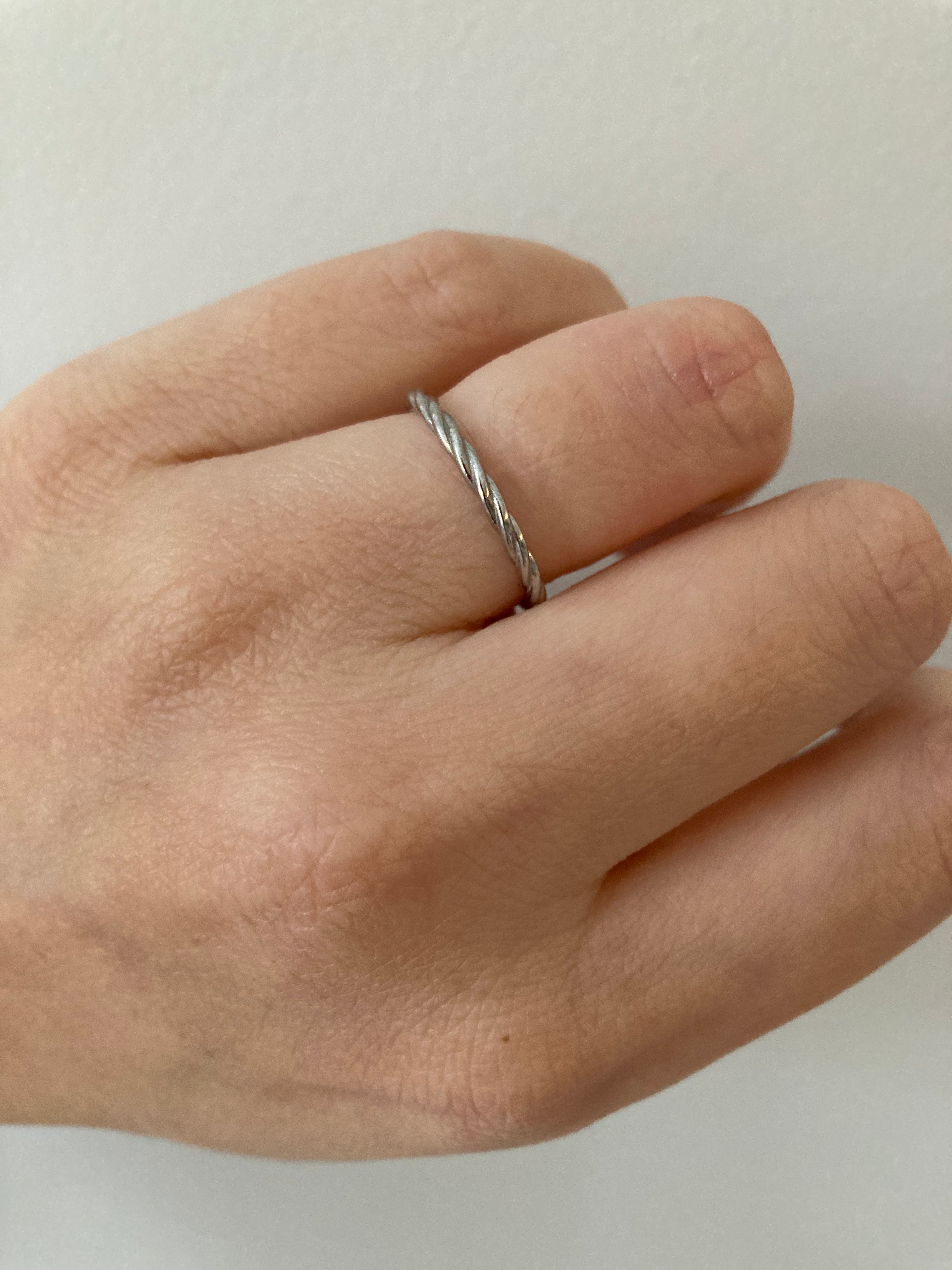 A white gold twisted rope ring worn on a person's finger agaisnt a neutral background.