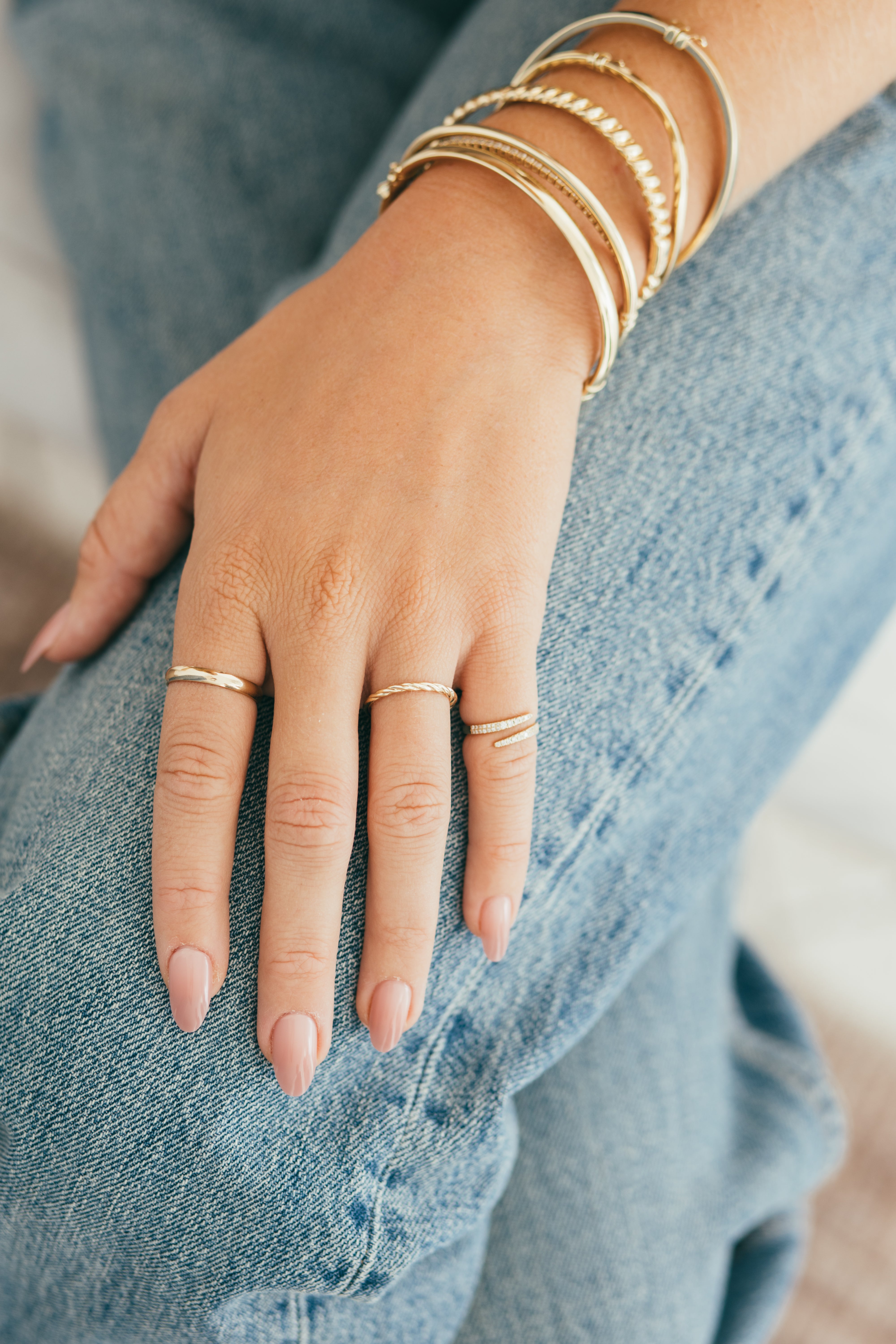 A woman's hand styled with gold rings and a stack of elegant gold bracelets set on denim jeans.