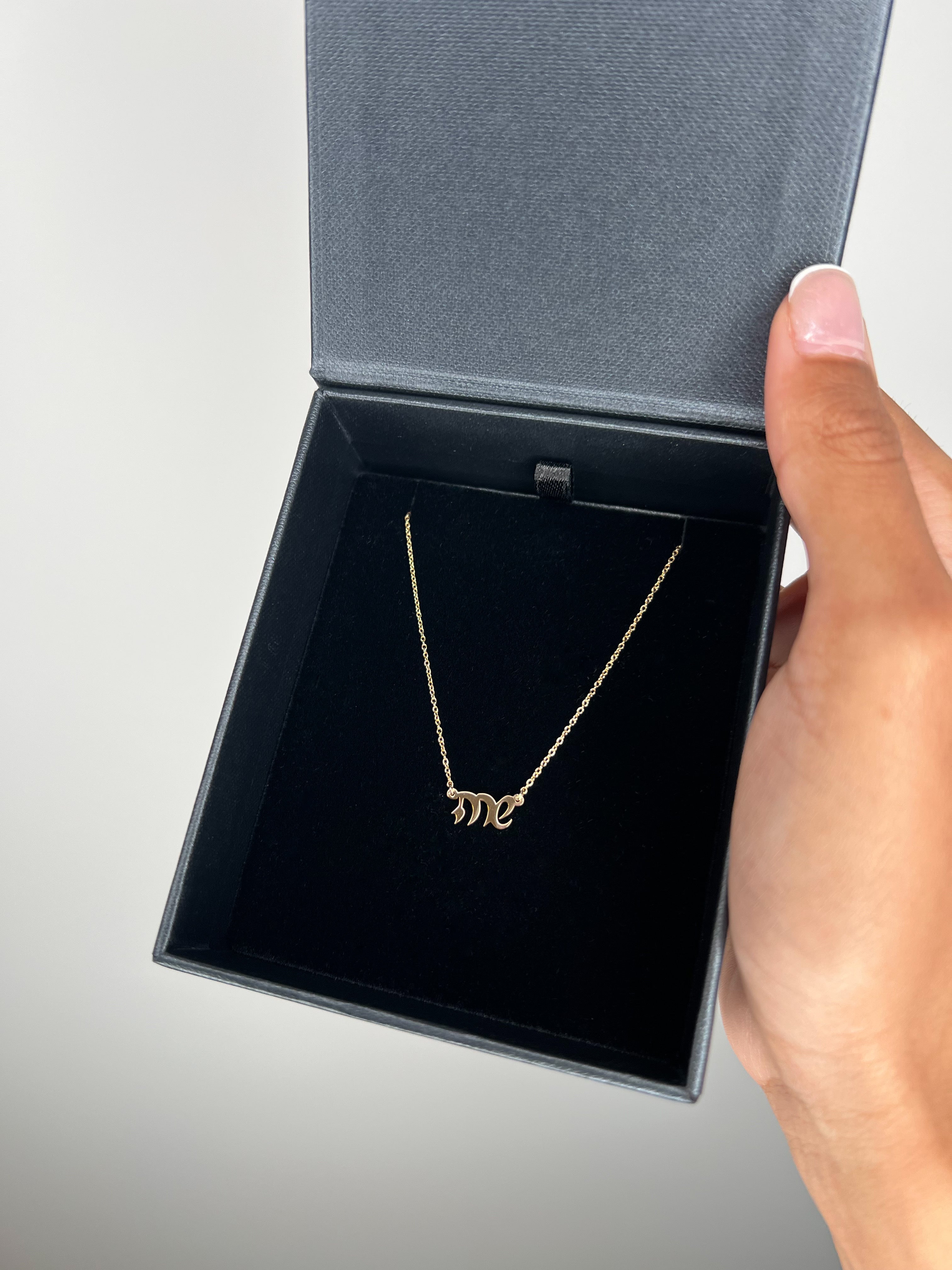 A gold name necklace displayed inside an open dark gray jewelry box, held by a hand against a neutral background.