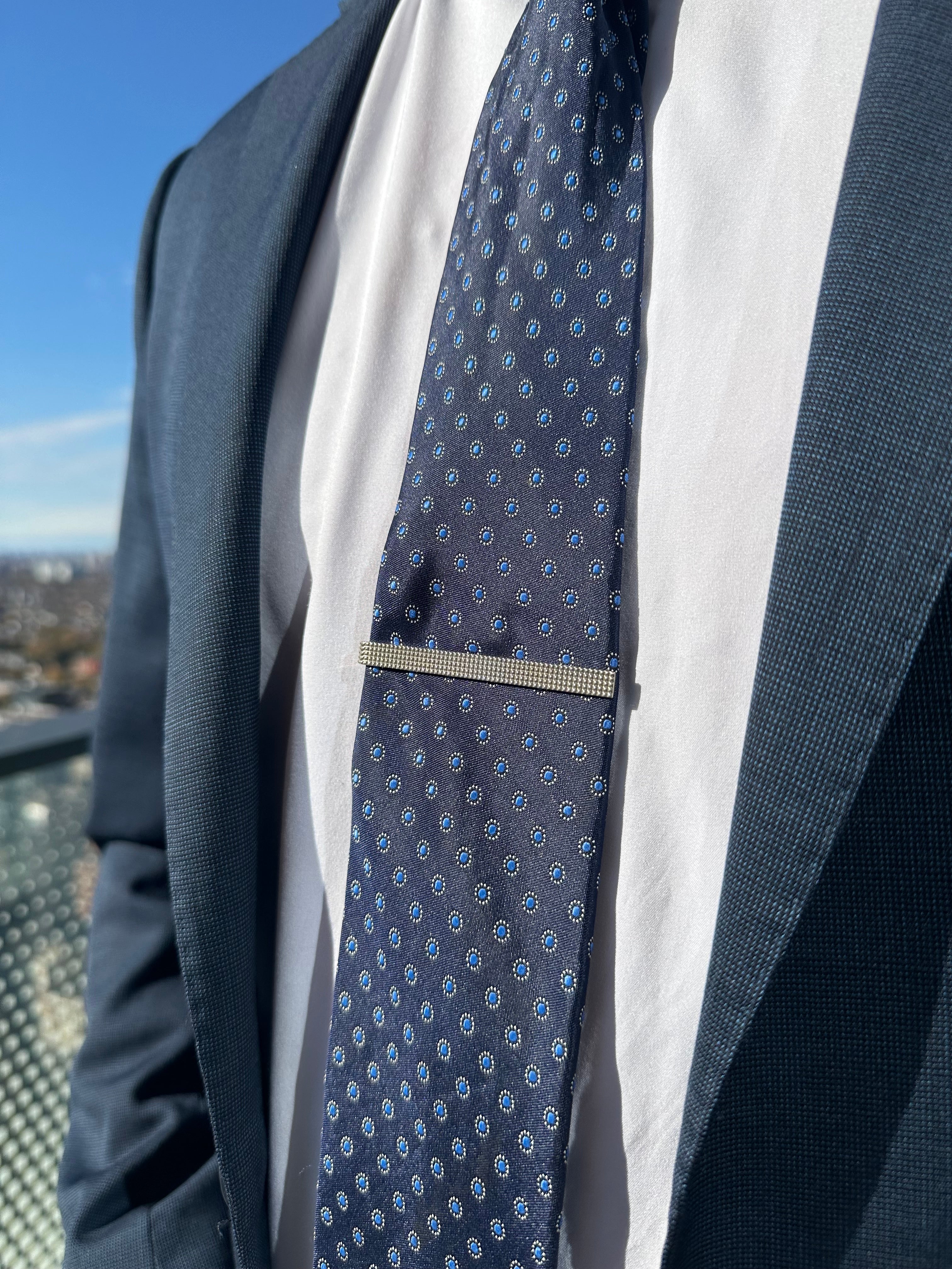A sterling silver textured tie bar worn on a men's dark blue patterned tie with a white dress shirt and dark blue suit jacket.