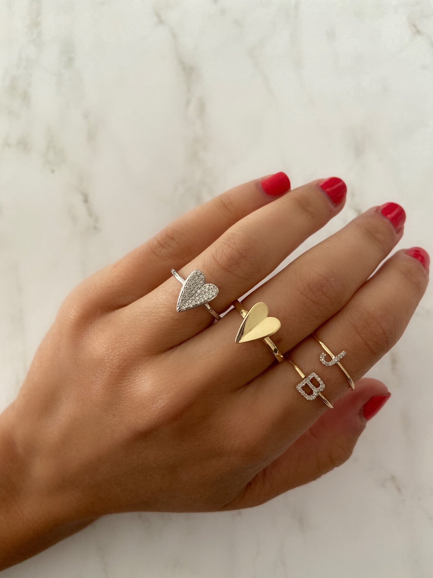 Gold heart rings styled on a person's hand with letter "J" and letter "B" rings with a white marble background