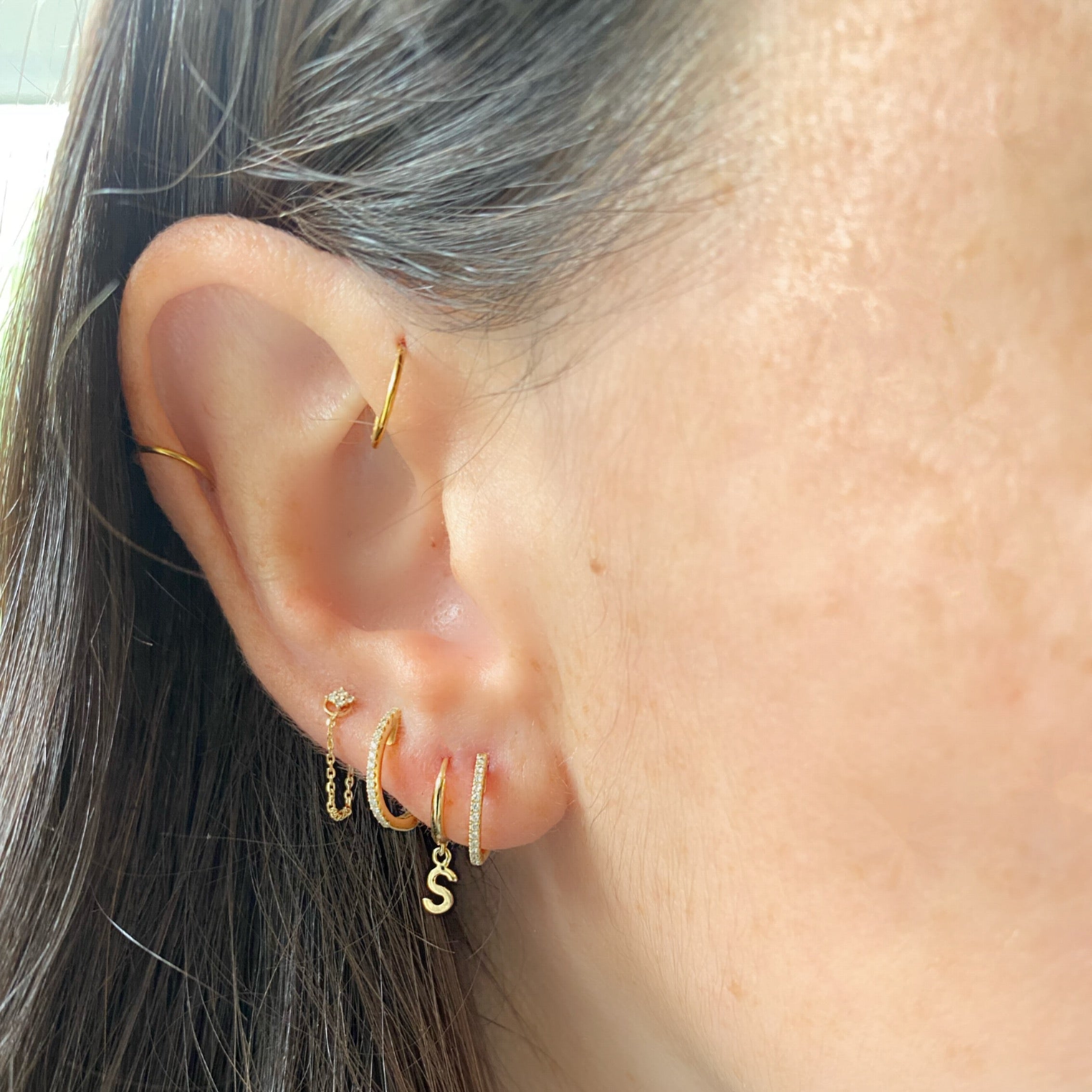 Close-up of an ear with gold hoop earrings and a 'S' charm.