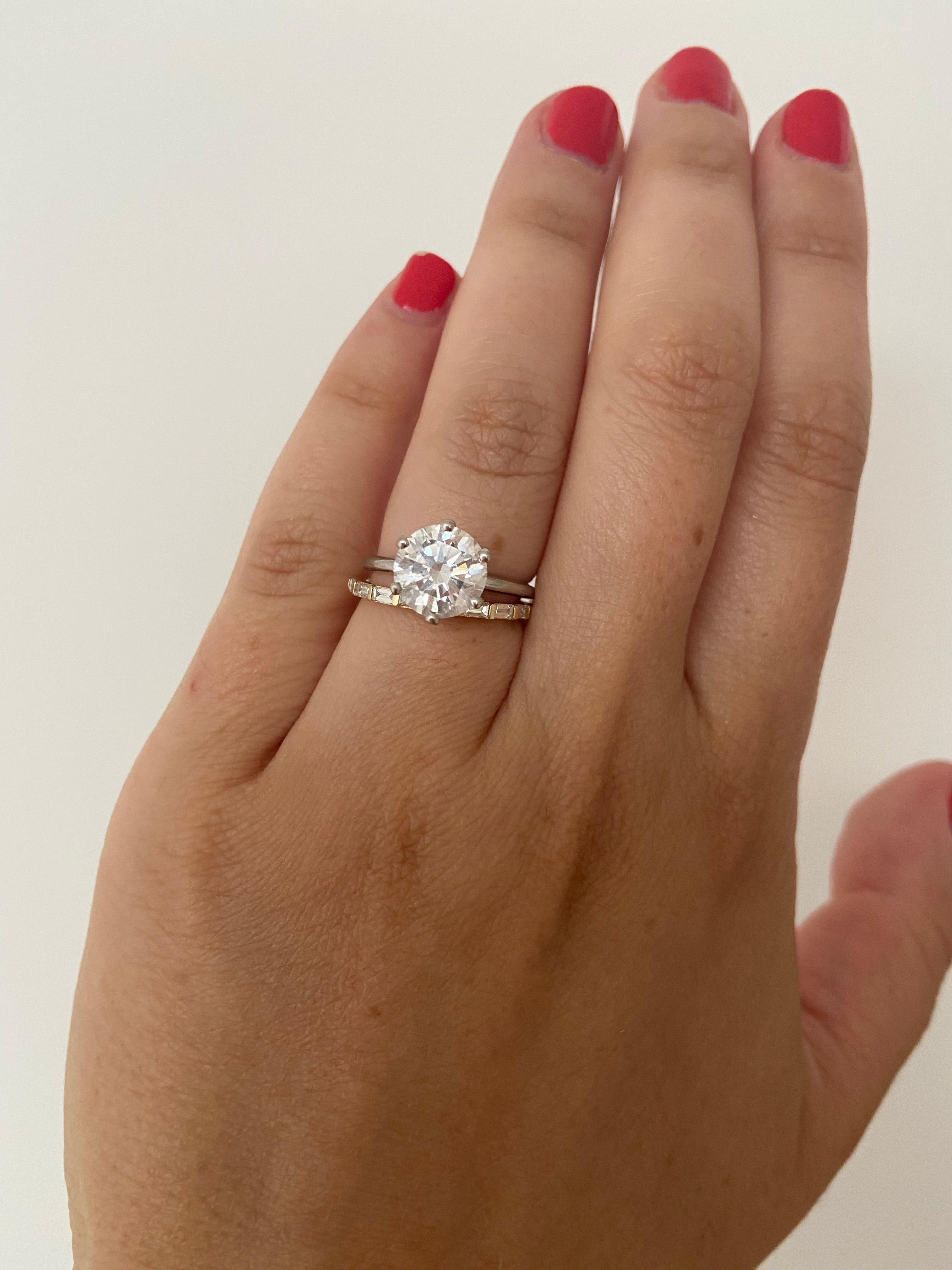 A woman's hand wearing a yellow gold diamond baguette ring stacked with a large round diamond ring on her index finger, on a neutral background.