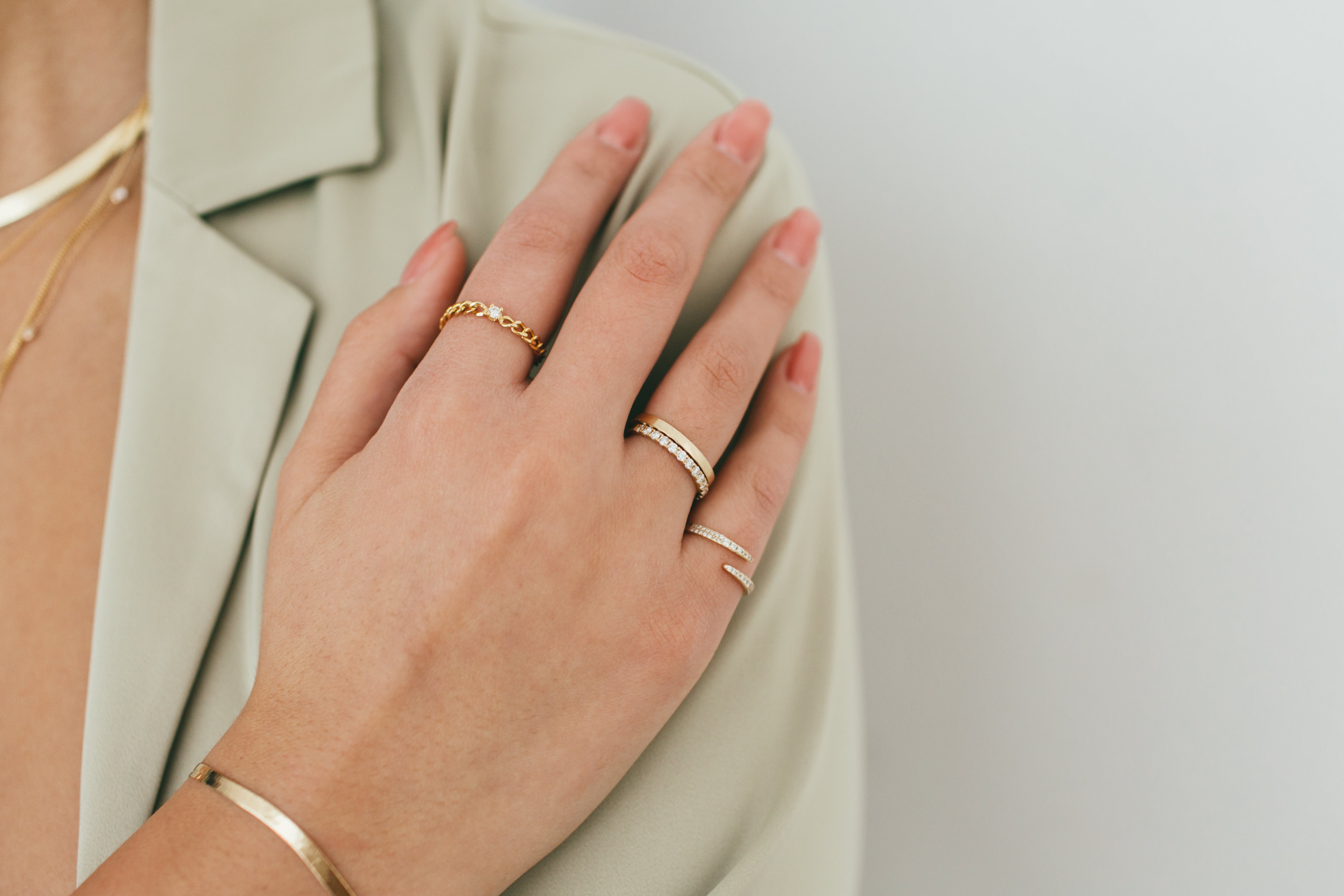 A woman's hand placed on her shoulder, styled with various gold and diamond rings and a flat gold bracelet.