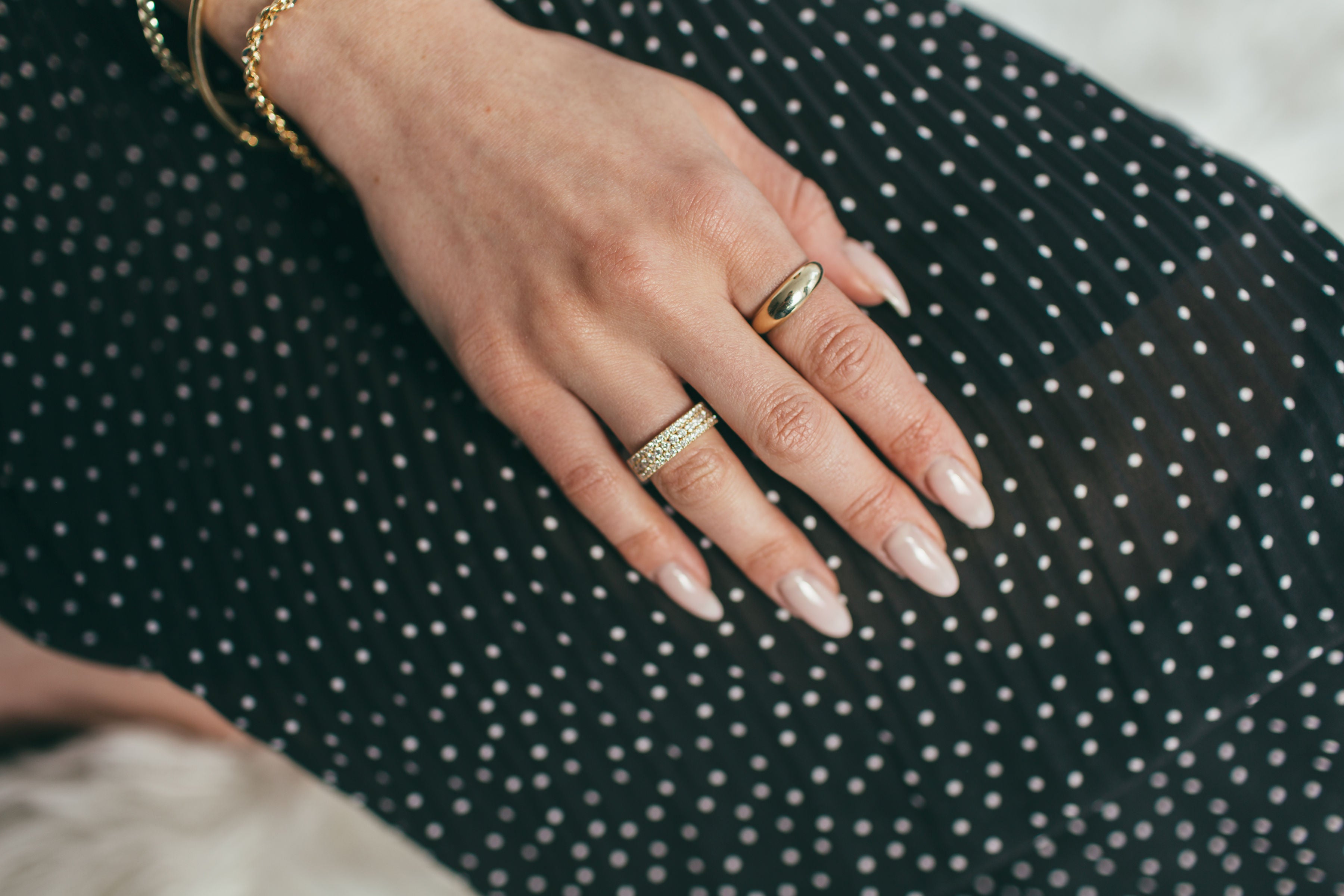 A close up of a woman's hand wearing a chunky yellow gold dome ring and a yellow gold large triple row pavé ring, resting on a black and white polka dot skirt.