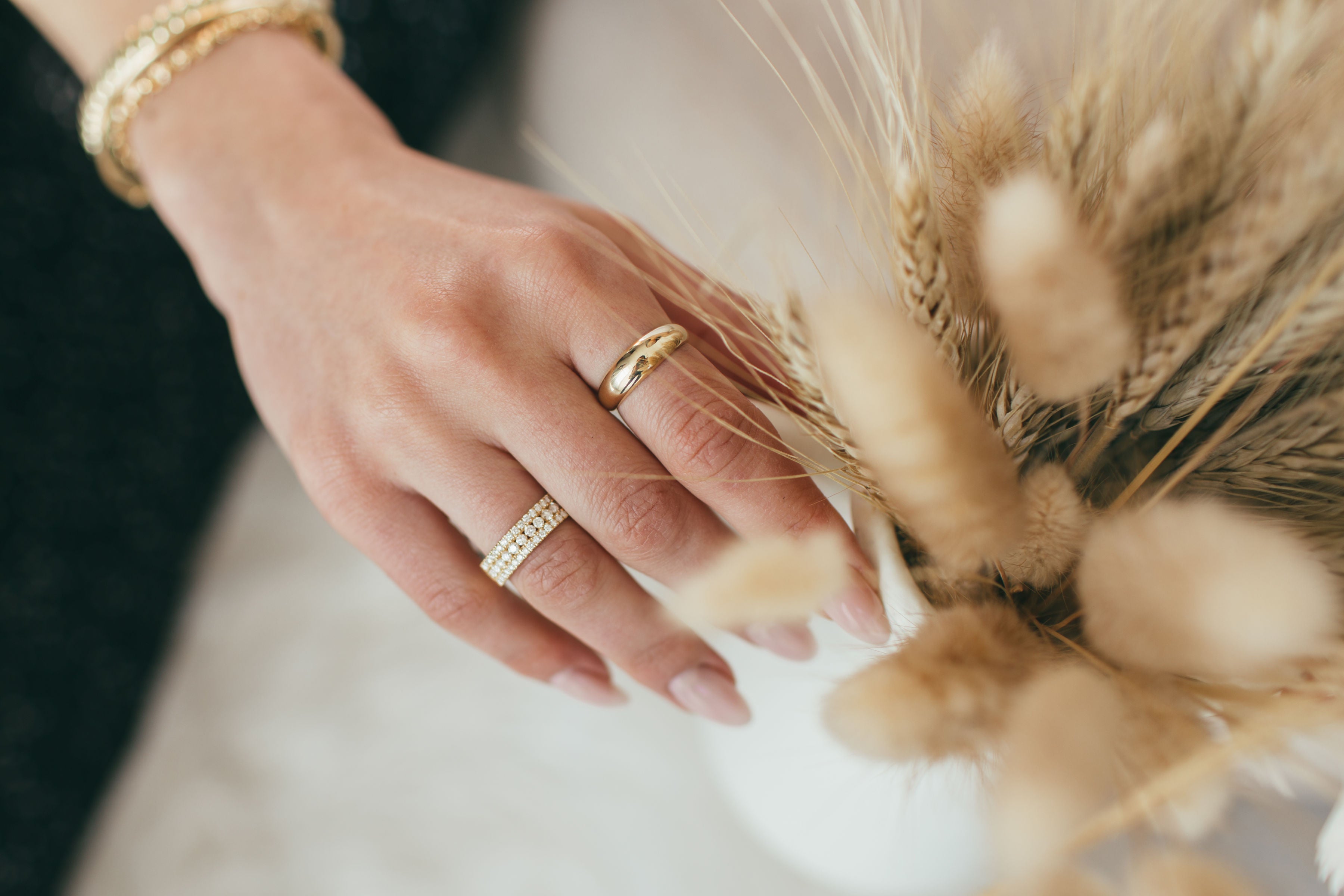 A close up of a woman's hand wearing a chunky yellow gold dome ring and a yellow gold large triple row pave ring, with blurred wheat stalks in the foreground and neutral colours in the background.