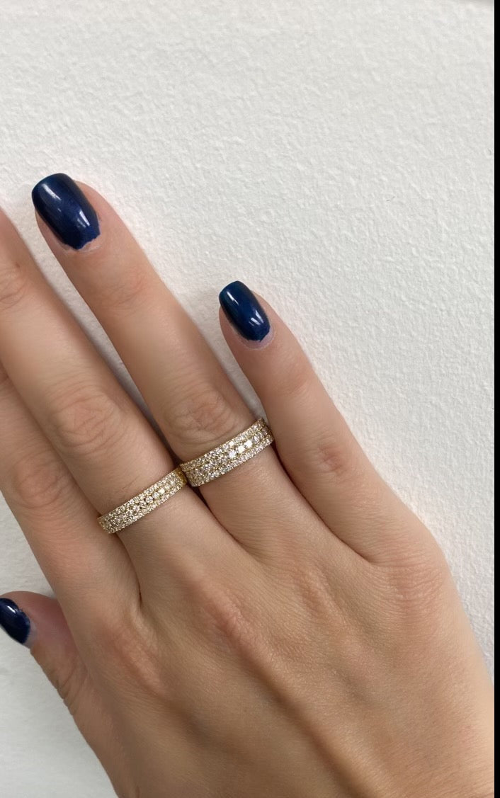 A close up of a woman's hand wearing a yellow gold large triple row pavé ring paired with a smaller triple row pavé ring, on a neutral background.