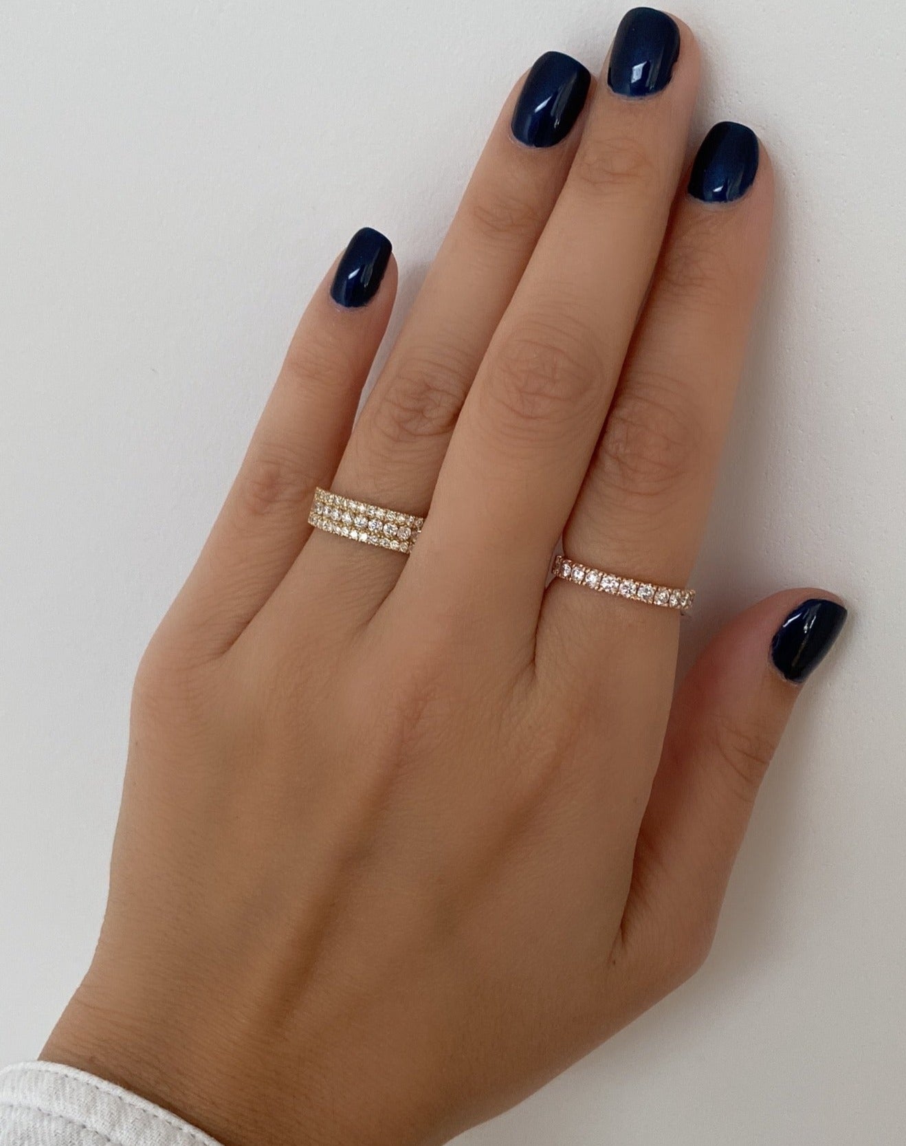 A close up a woman's hand wearing a yellow gold large triple row pavé ring on her index finger, and a thin diamond ring on her pointer finger, on a neutral background.