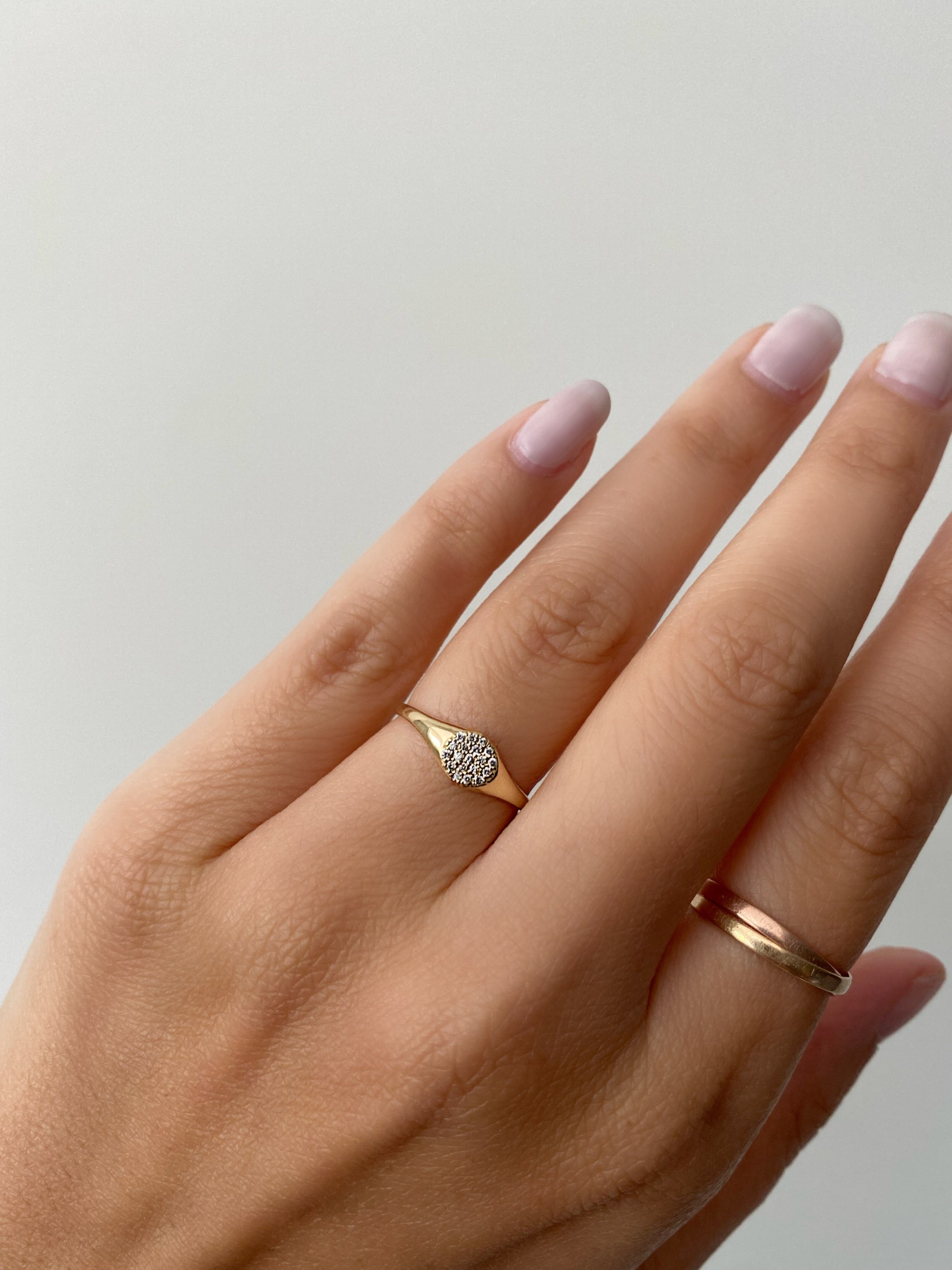 A close up of a woman's hand wearing a dazzling yellow gold mini diamond signet ring along with a simple gold wrap ring, on a neutral background.