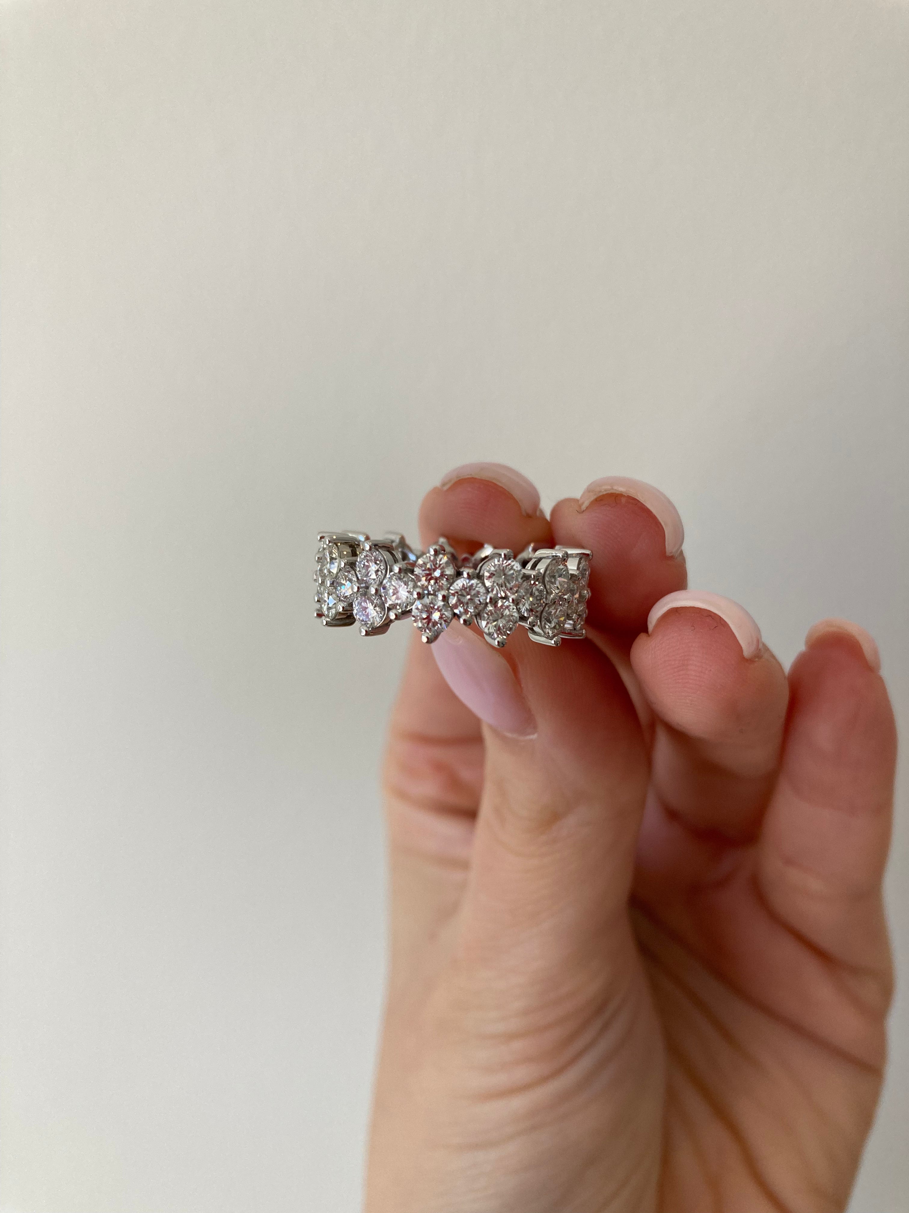 A close up of a white gold multi diamond eternity band, highlighting the striking diamonds, held between a person's fingers on a neutral background.